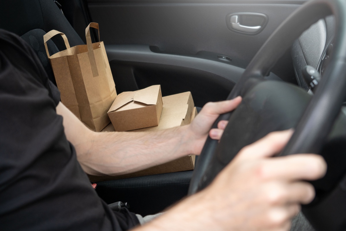 Takeout boxes and a paper bag on the passenger seat of a car while the driver holds the steering wheel