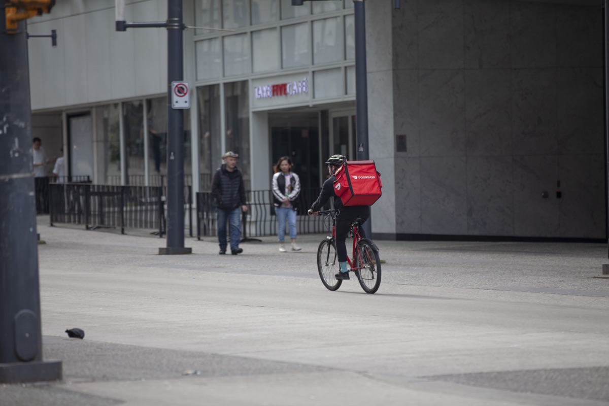 A bicycle courier with a red DoorDash insulated backpack rides across a downtown street