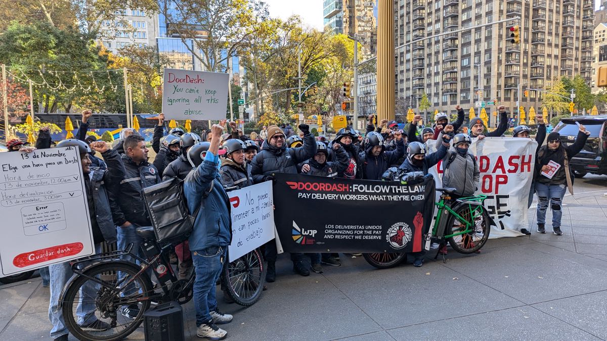 Delivery workers with bikes and helmets at a rally in New York City holding banners asking DoorDash to follow the law