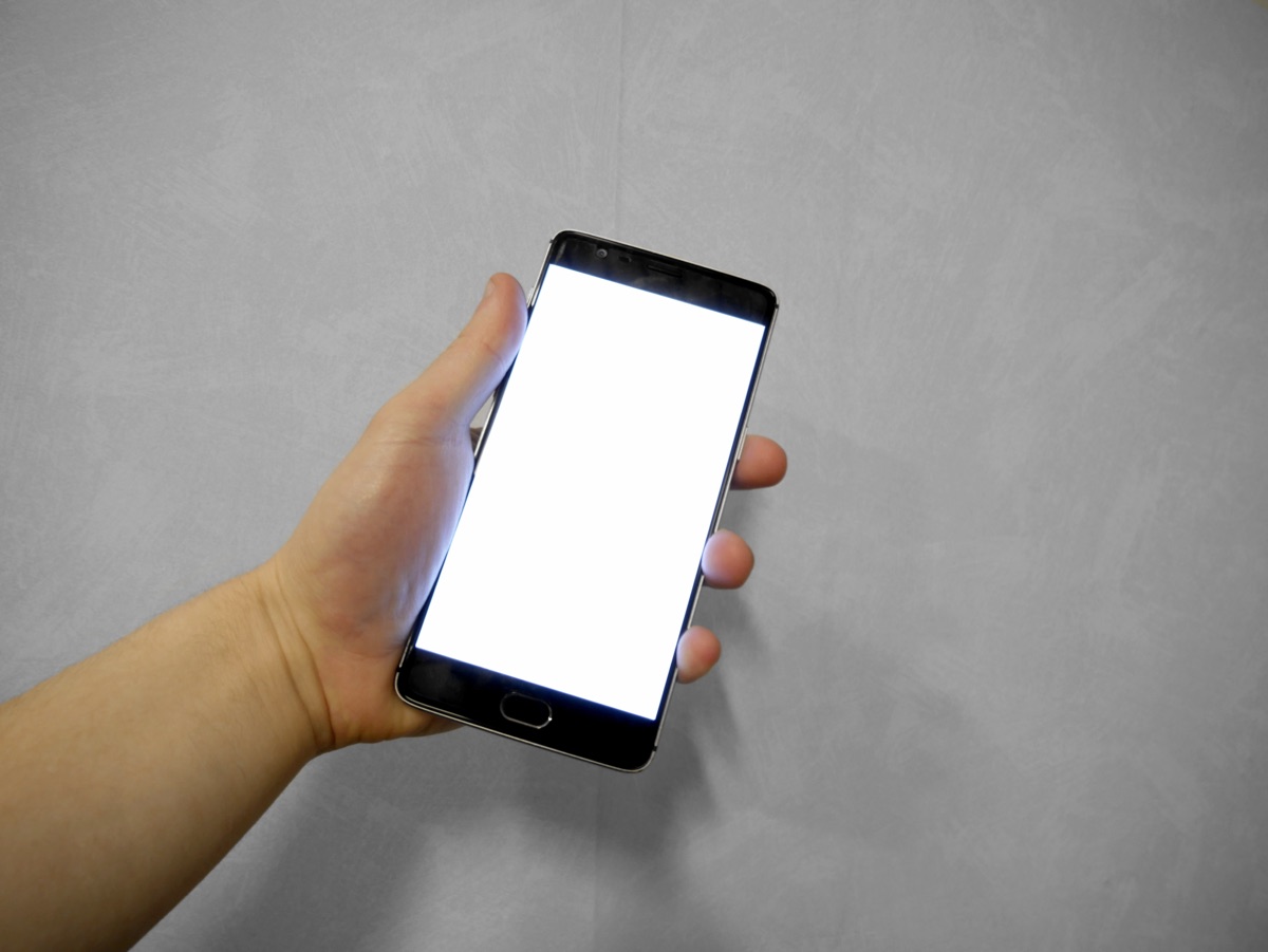 A hand holding a smartphone with a blank white screen against a gray wall