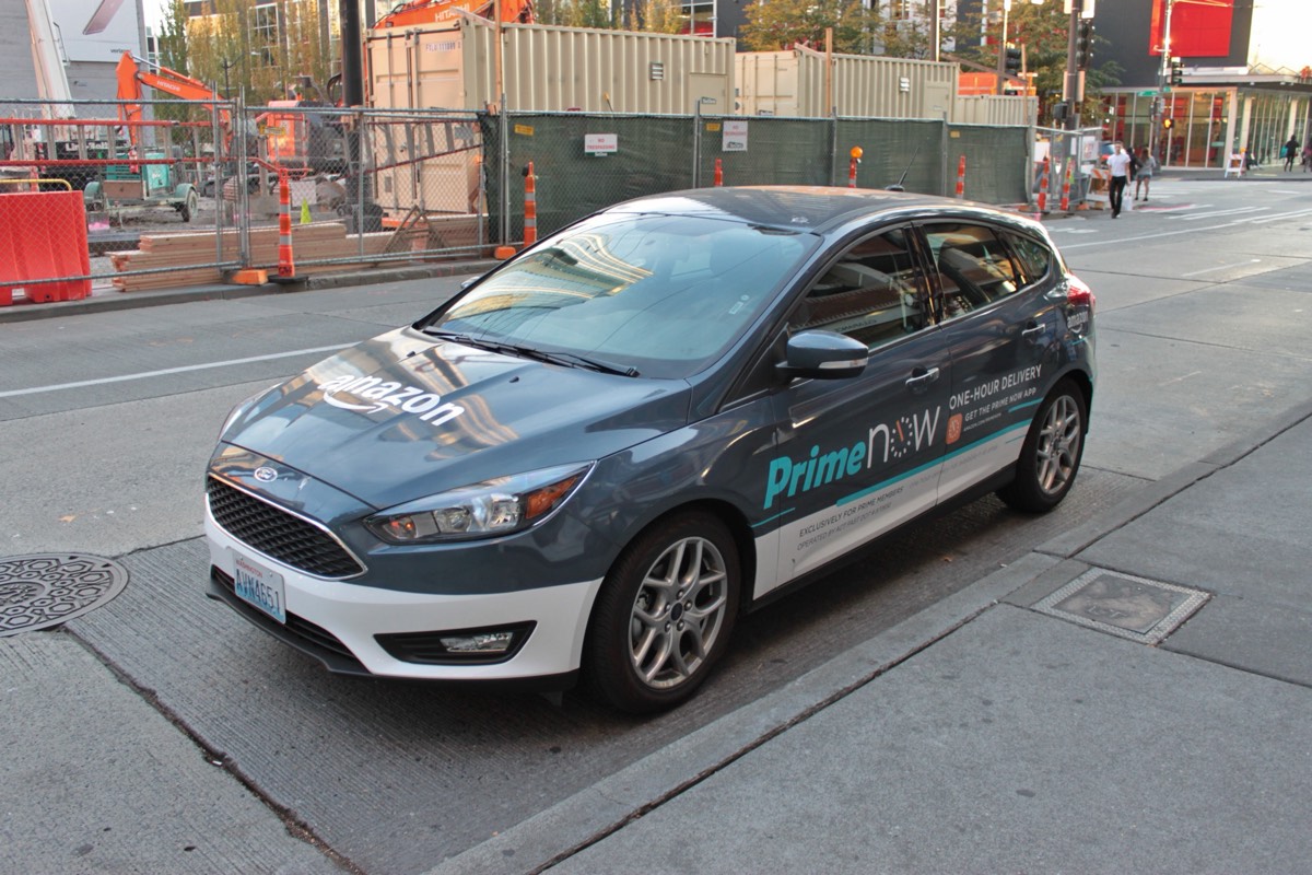 A blue hatchback with Amazon Prime Now delivery livery parked at a curb next to a construction site in Seattle