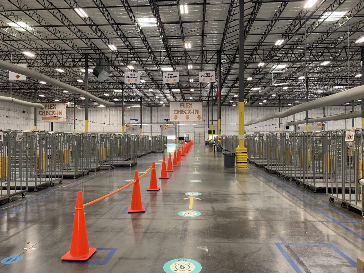 Inside an Amazon delivery station: a coned walkway between cart rows leads to signs reading Flex Check-In and Flex Checkout