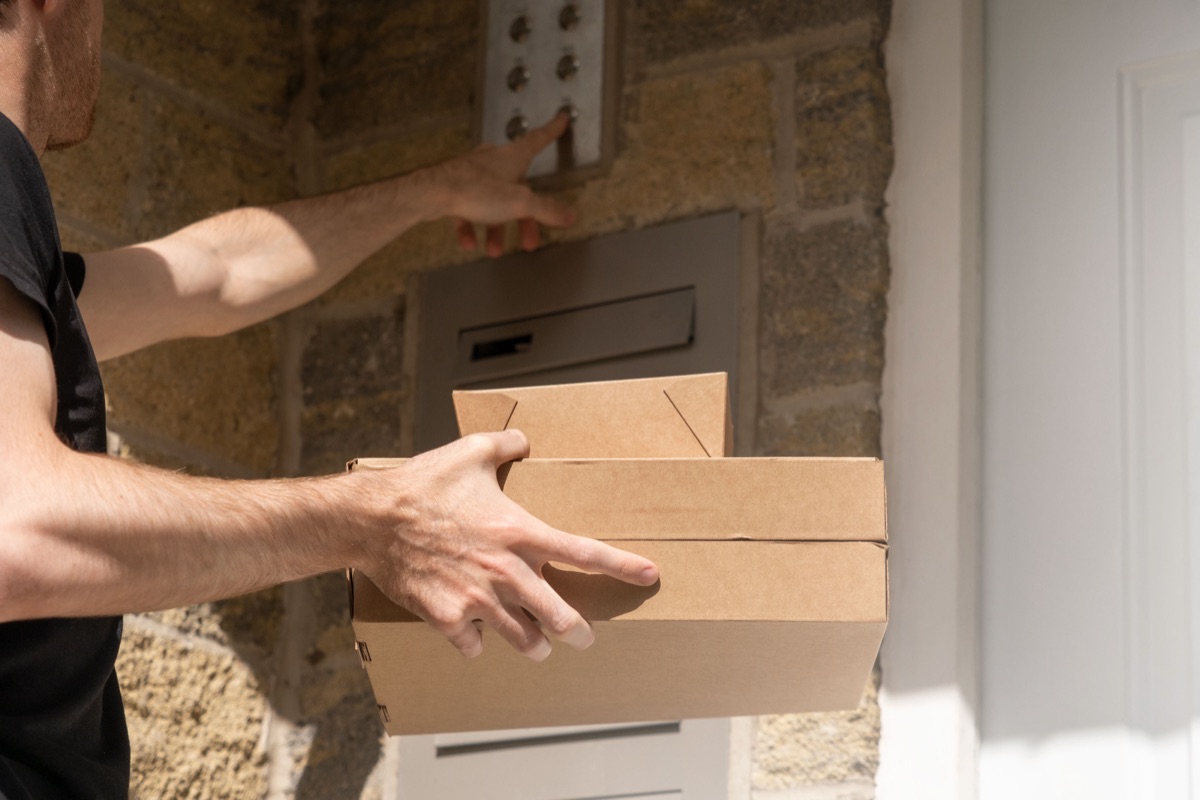 A delivery driver holding two takeout boxes reaches up to press an apartment building doorbell