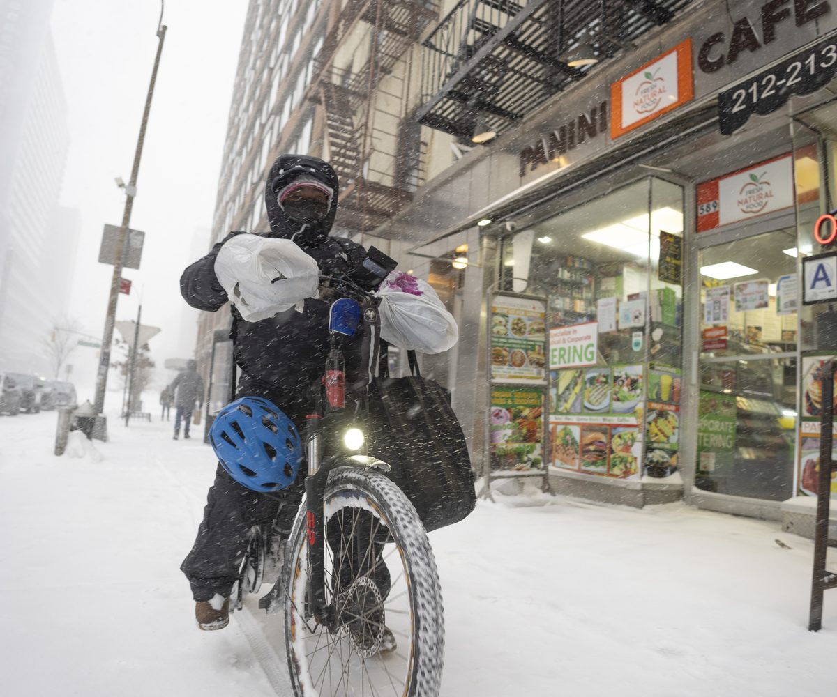 A food delivery courier on a bicycle with bags on the handlebars rides along a snow-covered Manhattan sidewalk